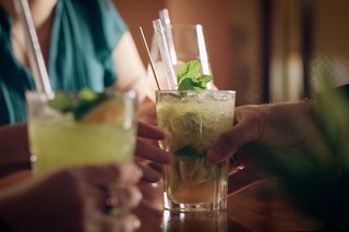 A woman in a crop top and wide-brimmed hat holds a colorful drink, with a sunny beach and palm trees in the background.