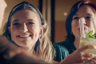 A bartender in an apron pours a cocktail from a shaker into a glass, surrounded by bottles and outdoor string lights.