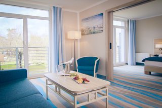 A smiling woman relaxes on a bed while a playful dog approaches her, creating a joyful and affectionate moment.