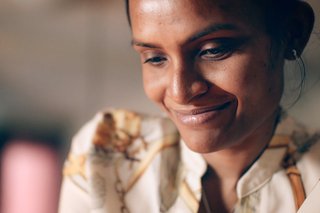 Two smiling women walk along a sunlit beach, one draped in a turquoise towel, wearing hats and swimwear, enjoying a carefree day by the ocean.