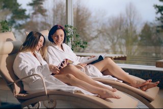 Two women in stylish outfits pose beside a vintage blue van labeled "PASÉA," with palm trees and modern architecture in the background.