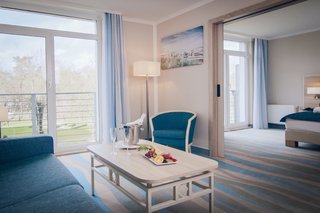 A bright, modern hotel room features a bed, couch, desk, and a scenic ocean view through large windows, with a guitar in the corner.