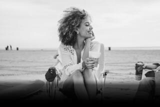 A smiling woman with curly hair enjoys a drink on the beach, radiating joy amidst a relaxed, social atmosphere.