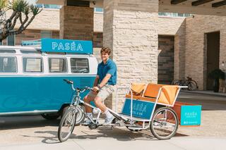A young man on a bicycle with an attached cart stands near a vintage blue van, promoting Paséa Hotel and Spa in a sunny location.