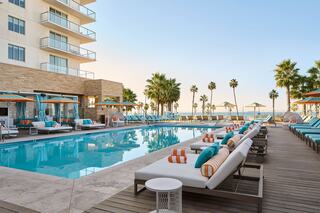 A stylish pool area features comfortable lounge chairs, cabanas, and palm trees, with a stunning ocean backdrop under a clear sky.
