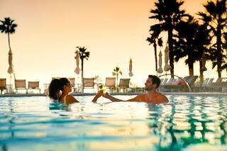 A couple enjoys drinks in a pool, surrounded by palm trees, under a vibrant sunset sky. Relaxation and connection shine in the moment.