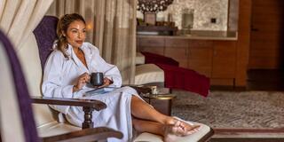 A relaxed woman in a robe sits comfortably in a luxurious setting, holding a cup while enjoying a moment of tranquility.