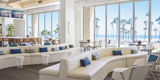 A bright, modern lounge features curved white seating with blue accents, large windows showcasing palm trees and ocean views.