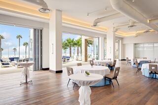 A spacious event venue features elegant tables, large windows with sea views, and palm trees creating a bright, inviting atmosphere.
