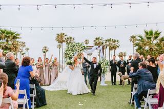 A joyous couple celebrates their wedding, surrounded by guests applauding, with palm trees and elegant decor in a beautiful outdoor venue.