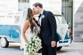 A bride and groom share a kiss, surrounded by a romantic atmosphere, with a vintage blue VW van in the background and a bouquet of white flowers.