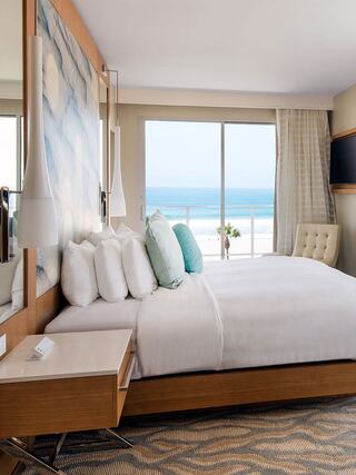 A serene hotel room features a large bed with pillows, modern decor, and a stunning ocean view through expansive windows.
