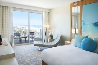 A modern hotel room features a balcony with coastal views, a chaise lounge, soft decor, and a cozy atmosphere ideal for relaxation.