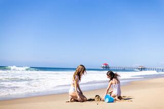 Two children play in the sand at a beach, building a castle as waves gently lap at the shore under a clear blue sky.