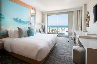 A modern hotel room featuring a large bed, vibrant wall art, and a stunning ocean view through large windows.