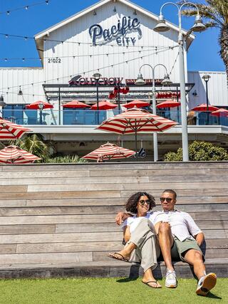 A couple relaxes on wooden steps amid vibrant umbrellas and palm trees, enjoying a sunny day at Ostsee City.