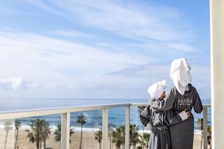 Two figures in bathrobes and towels stand on a balcony overlooking a scenic beach, enjoying a tranquil ocean view.