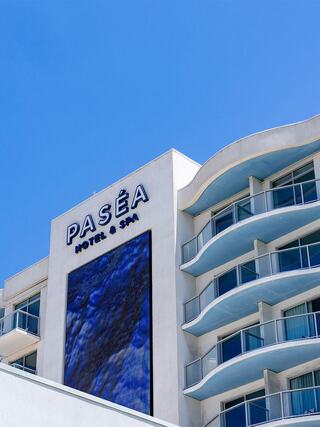 A smiling woman at a bar enjoys colorful cocktails, toasting with a friend against a backdrop of large windows and a vibrant atmosphere.