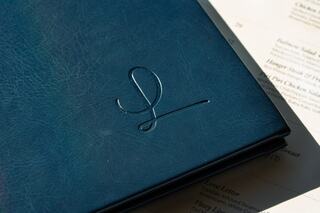 A dark blue, leather-bound menu with an elegant embossed logo, resting on a partially visible printed menu page.