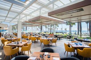 Bright and airy dining space with modern decor, featuring palm trees and ocean views. Cozy yellow chairs complement wooden tables.