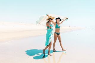 Two women in beachwear stroll along the shoreline, enjoying the sun and each other's company, with a clear blue sky overhead.