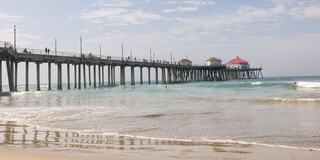 A seaside pier stretches over gentle waves, with people strolling along it and beachgoers enjoying the sun and surf under a clear sky.