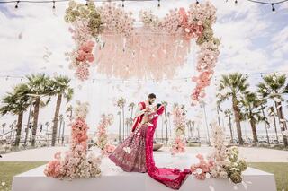 A couple embraces under a stunning floral arch adorned with pink and white blooms, surrounded by palm trees and a vibrant sky.