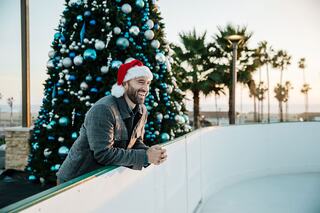 A man in a Santa hat leans against a rink railing, smiling near a festive Christmas tree, with palm trees and a sunset in the background.