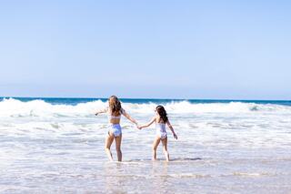 Two girls wade into the ocean, holding hands, as waves crash gently around them under a clear blue sky.