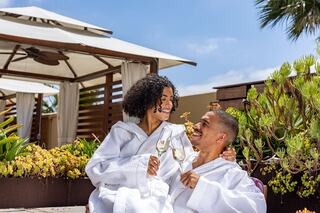 A couple in white robes smiles at each other while holding champagne glasses in a lush, sunny outdoor setting, surrounded by greenery.
