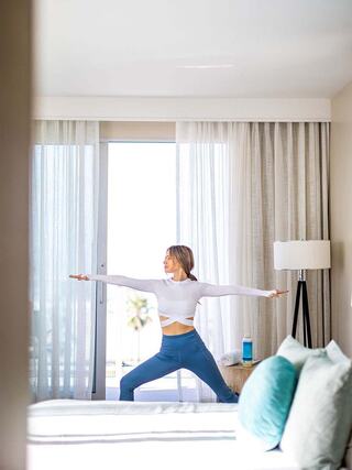 A woman practices yoga in a bright room, striking a warrior pose with large windows and soft furnishings in the background.