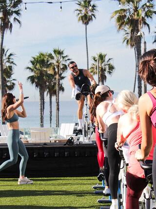 A group fitness class outdoors, featuring participants on stationary bikes with palm trees in the background. Motivation and energy are high.
