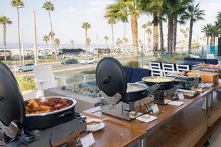 A vibrant outdoor buffet features trays of various dishes, with palm trees and a coastal view in the background.