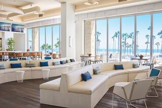 A bright, modern lounge features white furniture, blue accents, and large windows facing palm trees and the ocean, creating a serene atmosphere.