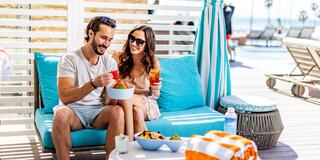 A cheerful couple enjoys snacks and drinks on a sunny day, relaxing on a cozy lounge with palm trees in the background.