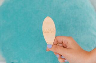 A hand holds a wooden key tag shaped like a surfboard, engraved with "Seehotel Grossherzog v. Mecklenburg," against a soft turquoise background.