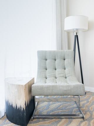 A modern gray armchair with tufted upholstery sits beside a unique wooden side table and a sleek floor lamp, all against a light backdrop.