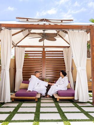 A couple in white robes relaxes on purple loungers under a stylish canopy, enjoying drinks in a serene outdoor setting.