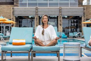 A woman meditates peacefully by a pool, surrounded by lounge chairs and vibrant umbrellas, exuding calm and relaxation.
