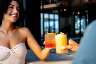 A woman smiles while toasting with colorful cocktails on a marble counter, set against a bright, modern backdrop.
