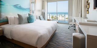 A spacious hotel room features a large bed, modern decor, and ocean views through expansive windows. Soft colors create a relaxing atmosphere.