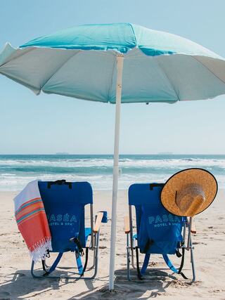 Two blue beach chairs with a straw sunhat and towel sit beneath a large umbrella, overlooking a serene sandy beach and the ocean.