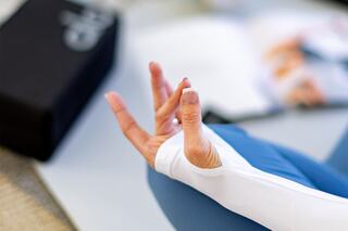 A hand in a meditative pose, dressed in a white long-sleeve top, with a serene backdrop including a yoga block and soft surfaces.