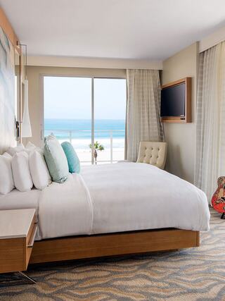 A bright, coastal bedroom features a large bed, ocean views through large windows, and modern decor, creating a serene atmosphere.