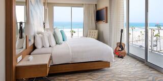 A modern, airy hotel room with a king-sized bed, sleek furniture, ocean view, and a guitar, creating a relaxing coastal atmosphere.