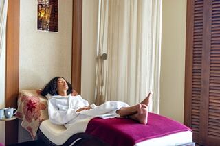 A woman relaxes in a cozy spa setting, wearing a robe, with a tranquil expression as sunlight filters through soft curtains.