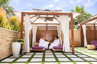 A couple relaxes in spa robes under a gazebo, enjoying drinks amidst a serene setting with lush greenery and soft, sunny skies.