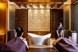 Two massage therapists work in a serene spa setting, featuring an elegant bathtub and an intricately designed wall panel.