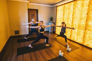 Three women practice yoga in a serene indoor space, performing warrior poses on mats with a warm, glowing wall in the background.