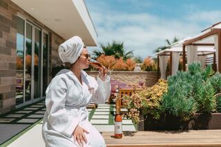 A woman in a bathrobe sips wine on a sunny patio, surrounded by greenery and modern outdoor furniture, exuding relaxation and luxury.